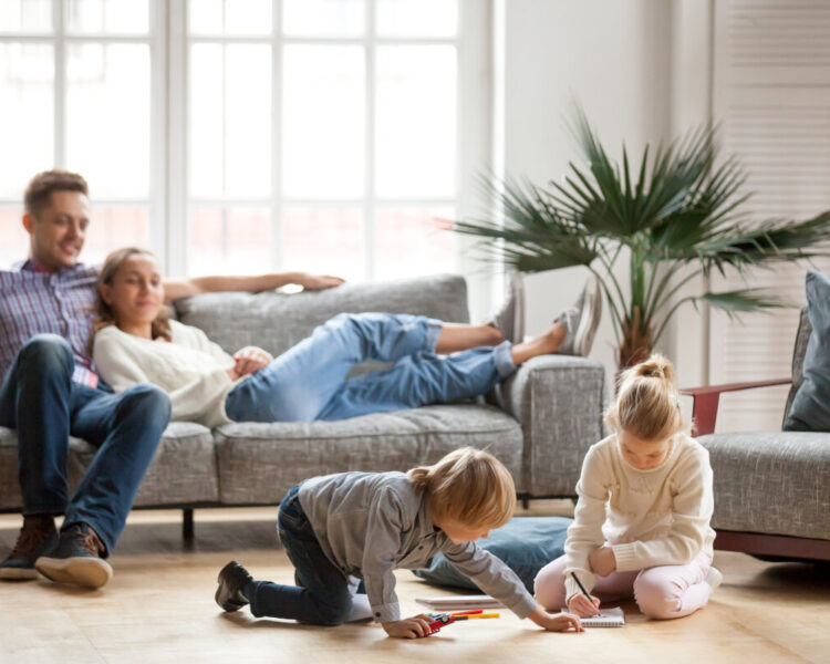 Children sister and brother playing drawing together on floor while young parents relaxing at home on sofa, little boy girl having fun, friendship between siblings, family leisure time in living room